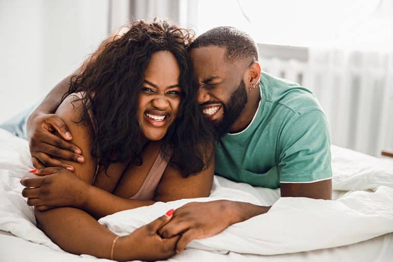 A joyful couple lies on a bed, smiling and hugging each other closely, enjoying a happy moment of autumn romance together in a bright, cozy room.