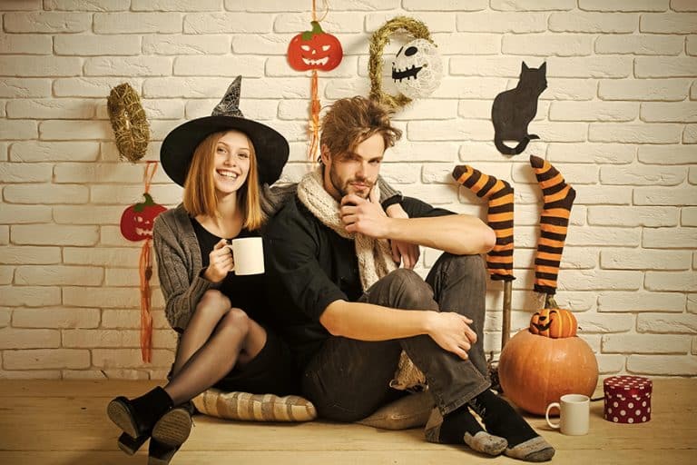 Two people in spooky and sexy Halloween costumes sit against a white brick wall decorated with pumpkins, a black cat cutout, and striped witch legs. One wears a witch hat and smiles; both hold mugs. A festive, cozy Halloween scene.