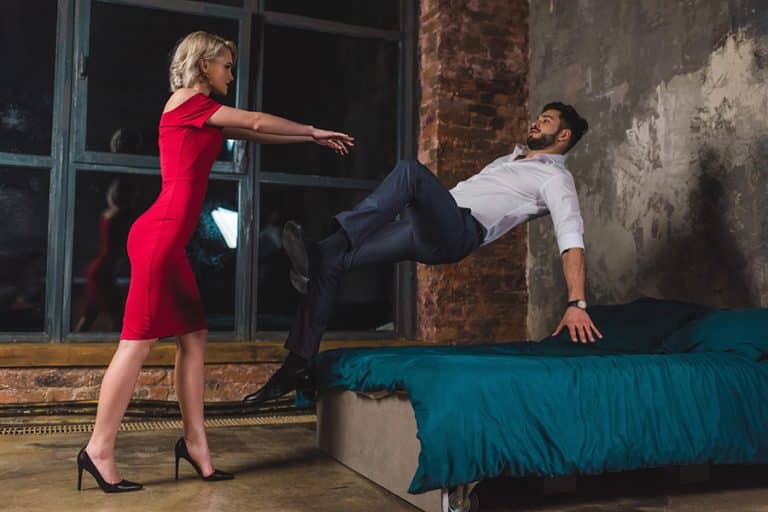 A woman in a red dress, intent on reigniting passion, pushes a man in a white shirt and dark pants onto a bed in an industrial-style room with brick walls and large windows.
