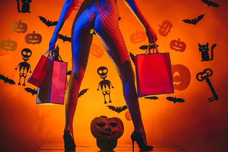A person in fishnet stockings and high heels holds shopping bags, standing in front of Halloween-themed decorations with bats, skeletons, and pumpkins. Channeling a Trick or Tease vibe, they strike a confident pose beside a carved jack-o-lantern sitting on the floor.